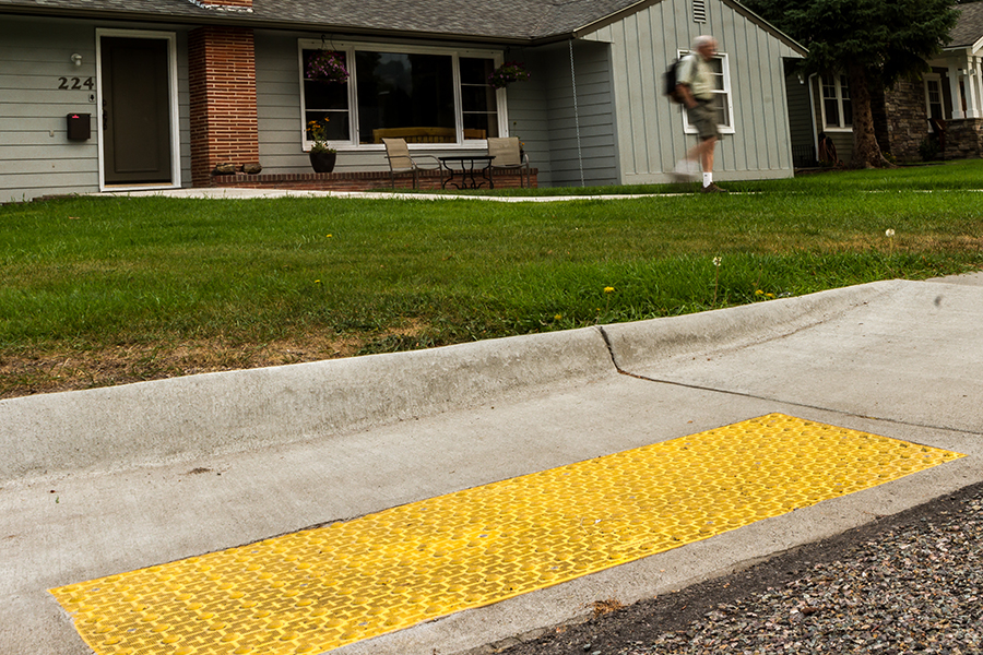 A zoomed in picture showing a details of a curb-ramp from a sidewalk to a street. The ramp has a yellow plate with dome-shaped bumps that act as a tactile warning to pedestrians. A fellow with shorts and a backpack strolls away from the house in the background.
